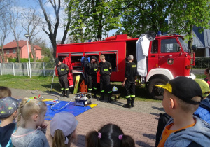 Dzieci stoją na dworze wokół wozu strażackiego, strażacy prezentują narzędzia znajdujące się w wozie strażackim..